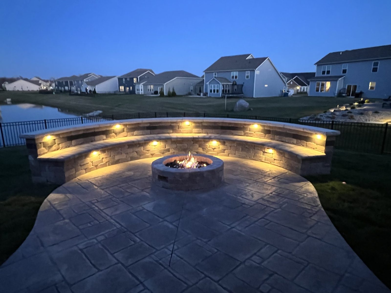 Stamped concrete patio with a curved sitting wall, landscape lighting and a fire pit at dusk