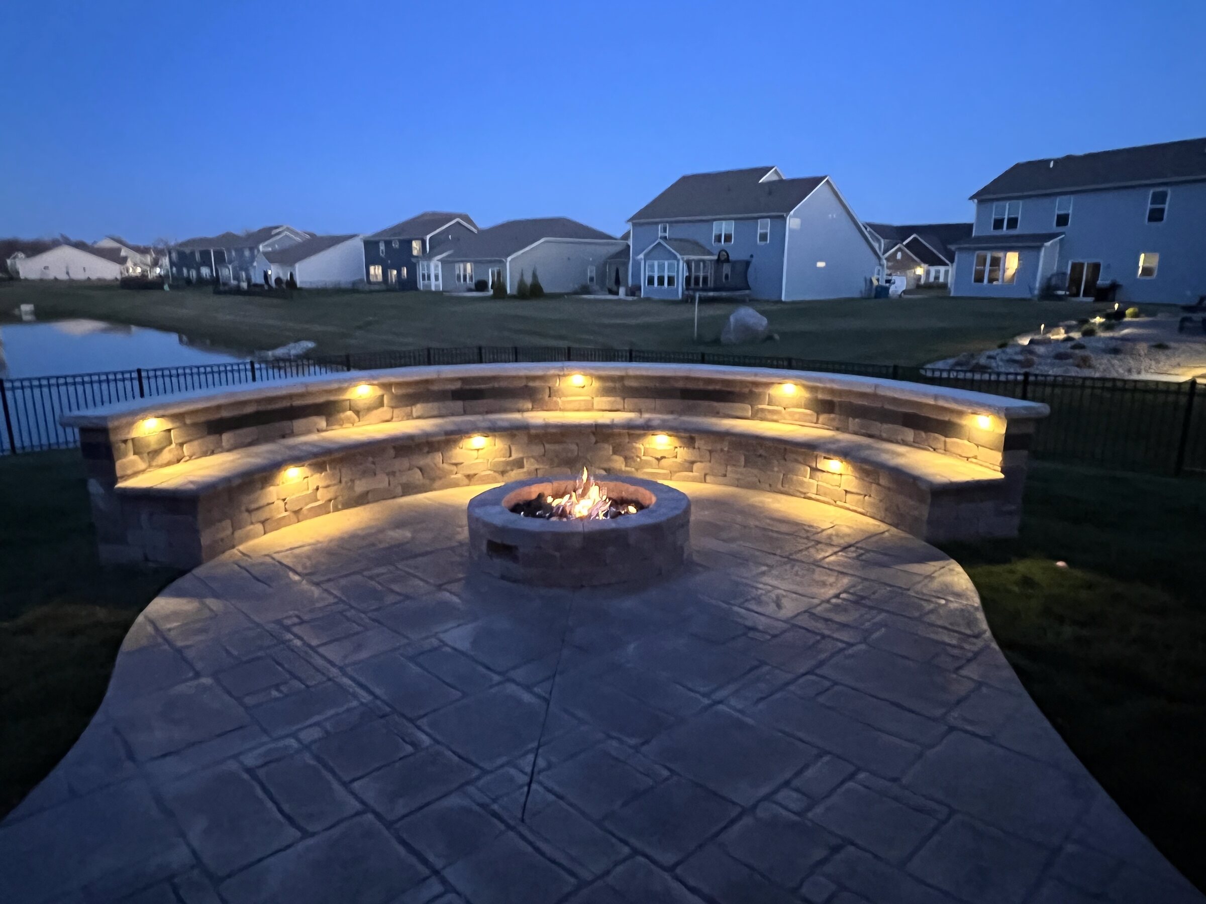 Stamped concrete patio with a curved sitting wall, landscape lighting and a fire pit at dusk